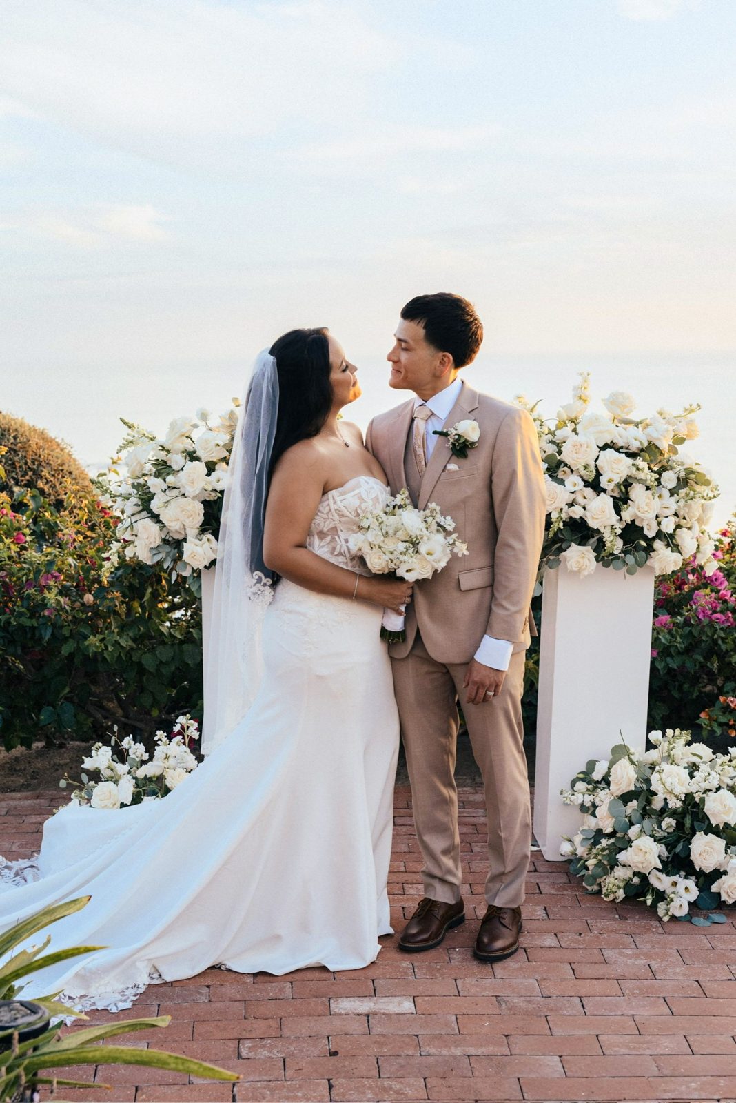 Newlyweds between white floral pillars on an ocean-view terrace